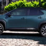 2025 Toyota Yaris Cross zijprofiel met verhoogde SUV-houding, zwarte carrosseriebescherming, lichtmetalen velgen op kasseistraat met Europese architectuur