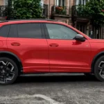 Zijaanzicht van rode 2025 Volkswagen Tiguan geparkeerd op kinderkopjes met compleet SUV-profiel, lichtmetalen wielen en stedelijke achtergrond