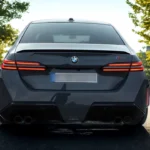 Direct achteraanzicht van 2025 BMW M5 met quad uitlaatpijpen, M5-badge, BMW-logo en volle breedte achterlichten op donkere sedan