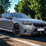 Voor driekwart aanzicht van 2025 BMW M5 in stedelijke omgeving met gebouwen en bomen, sportieve voorbumper en slanke vormgeving tonend