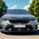 Vooraanzicht van 2025 BMW M5 luxe sedan in parkomgeving met bomen en bloemen, volledige voorkant en elegante vormgeving tonend