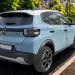 Achteraanzicht van lichtblauwe 2025 Citroën ë-C3 elektrische auto op stadsstraat met gebouwen en mensen op achtergrond, LED-achterlichten, achterspoiler en ë-C3 elektrische badges tonend