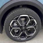 Gedetailleerd beeld van 2025 Citroën ë-C3 lichtmetalen velg met uniek hexagonaal geometrisch patroon, zwarte en bewerkte afwerking, gemonteerd op lichtblauwe auto met zwarte wielkasten bekleding