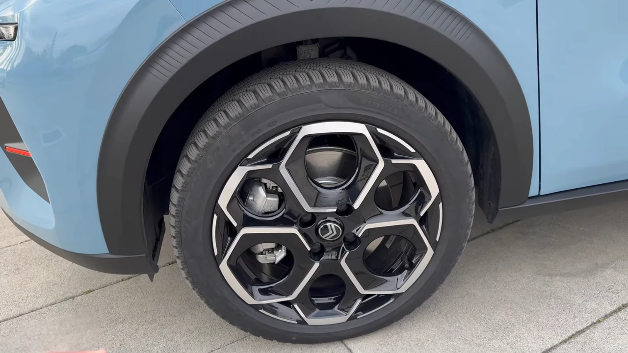 Gedetailleerd beeld van 2025 Citroën ë-C3 lichtmetalen velg met uniek hexagonaal geometrisch patroon, zwarte en bewerkte afwerking, gemonteerd op lichtblauwe auto met zwarte wielkasten bekleding