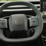 2025 Citroën ë-C3 stuurwiel met geïntegreerde bedieningsknoppen aan beide zijden, Citroën chevron-logo in het midden, grijze interieurafwerking en modern stuurwieldesign met touchscreen op achtergrond