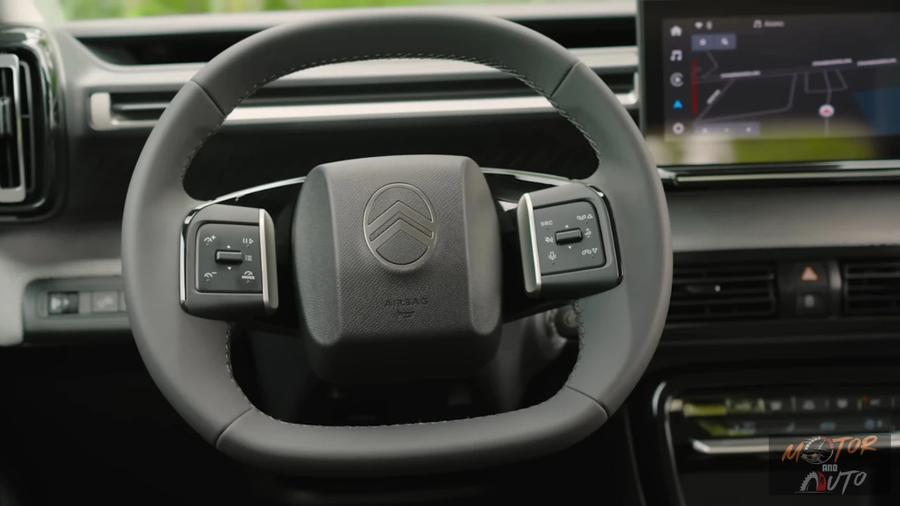 2025 Citroën ë-C3 stuurwiel met geïntegreerde bedieningsknoppen aan beide zijden, Citroën chevron-logo in het midden, grijze interieurafwerking en modern stuurwieldesign met touchscreen op achtergrond