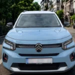 Vooraanzicht van lichtblauwe 2025 Citroën ë-C3 elektrische auto op Europese stadsstraat met historische gebouwen op achtergrond, LED-koplampen, chevron-grille en compacte crossover-proporties tonend