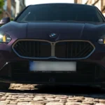 2025 BMW 1 Serie vooraanzicht close-up op kasseienstraat met verlichte LED-koplampen, prominente BMW niergrille en stedelijke architectuur op de achtergrond in paarse metallic afwerking