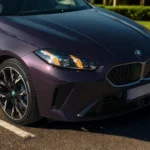 2025 BMW 1 Serie zijdetail met sportieve lichtmetalen velgen met blauwe remklauwen, gebeeldhouwde carrosseriepanelen en premium paarse metallic lak in natuurlijk licht