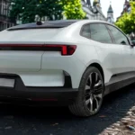 Achter-driekwart aanzicht van witte 2025 Polestar 4 elektrische auto geparkeerd op kinderkopjesstraat met traditionele Europese architectuur op de achtergrond, toont fastback-ontwerp en LED-achterlichten