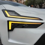 Close-up van de voorkoplamp van de 2025 Polestar 4 met LED-dagrijverlichting met gele amber accenten en modern hoekig ontwerp op witte voertuigcarrosserie