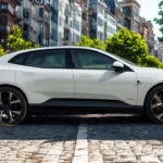 Zijprofiel van witte 2025 Polestar 4 op kinderkopjesstraat met stedelijke gebouwen op de achtergrond, toont de coupé-SUV-verhoudingen van het voertuig en zwart wielontwerp