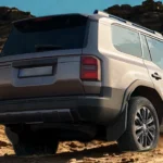 Achter driekwart aanzicht van witte 2025 Toyota Land Cruiser SUV klimmend over rotsachtig woestijnterrein met bergen op de achtergrond