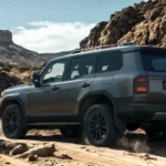 Achter driekwart aanzicht van donkergrijze 2025 Toyota Land Cruiser klimmend over grote rotsen in woestijnlandschap met bergen zichtbaar in de verte