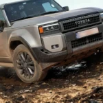 Voor driekwart hoek van beige 2025 Toyota Land Cruiser klimmend op steile rotsachtige helling met stofwolken, demonstrerend off-road prestatie