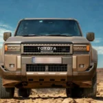 Direct vooraanzicht van beige 2025 Toyota Land Cruiser met kenmerkende grille, TOYOTA letters, en LED koplampen gepositioneerd in zanderige woestijn omgeving