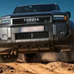 Vooraanzicht van 2025 Toyota Land Cruiser rijdend door zanderig woestijnterrein met stofwolken, met prominente Toyota grille en LED koplamp design