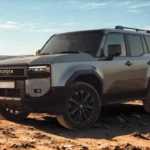 Zijprofiel van grijze 2025 Toyota Land Cruiser gepositioneerd op woestijnterrein met dramatische zonsondergang verlichting en berg silhouetten op achtergrond