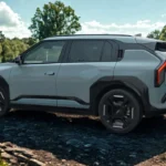 Volledig zijaanzicht van lichtgroene 2025 Kia EV3 elektrische SUV met zwarte lichtmetalen velgen geparkeerd in park met weelderige groene bomen en bewolkte hemel