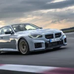 Zijaanzicht van 2025 BMW M2 CS in beweging met koolstofvezel dak, verlaagde houding, Gold Bronze gesmede wielen, circuitbanden, en aggressieve aerodynamische zijkant elementen