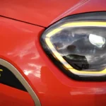 Gedetailleerd zicht van Mini Aceman hoekige LED-koplampbehuizing met verlichte gele DRL-ring, hoofd-LED-projector en gele "S" elektrische aanduiding badge op rode carrosserie