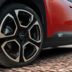 Gedetailleerd zicht van Mini Aceman lichtmetalen velg met geometrisch spakenontwerp, Mini-naafkap, zwarte en zilveren afwerking, gemonteerd op rode elektrische crossover met beschermende carrosseriebescherming