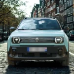 Vooraanzicht van de lichtblauwe 2025 Renault 4 E-Tech Electric op een kasseienstraat met kenmerkende LED-koplampen en verlicht Renault-logo