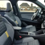 Interieurzicht van de voorstoelen van de 2025 Renault 4 E-Tech Electric met doorgestikt patroon, stoffen bekleding met gele accentnaden en opbergruimte in de middenconsole
