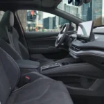Zijaanzicht van 2025 Škoda Enyaq interieur met bestuurders- en passagiersstoelen in grijze stofbekleding, centraal infotainment scherm en modern dashboard ontwerp met stadszicht door ramen