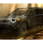 2026 Jeep Cherokee hybride SUV in grijze kleur rijdend door bospad met stofwolken, zijprofiel aanzicht met verhoogde bodemvrijheid en avontuur-klaar ontwerp in natuurlijke verlichting