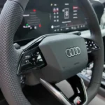 Gedetailleerd zicht op het stuurwiel van de 2026 Audi A6 met touch-gevoelige bedieningspanelen, Audi-logo, schakelpeddels en digitaal instrumentencluster op de achtergrond