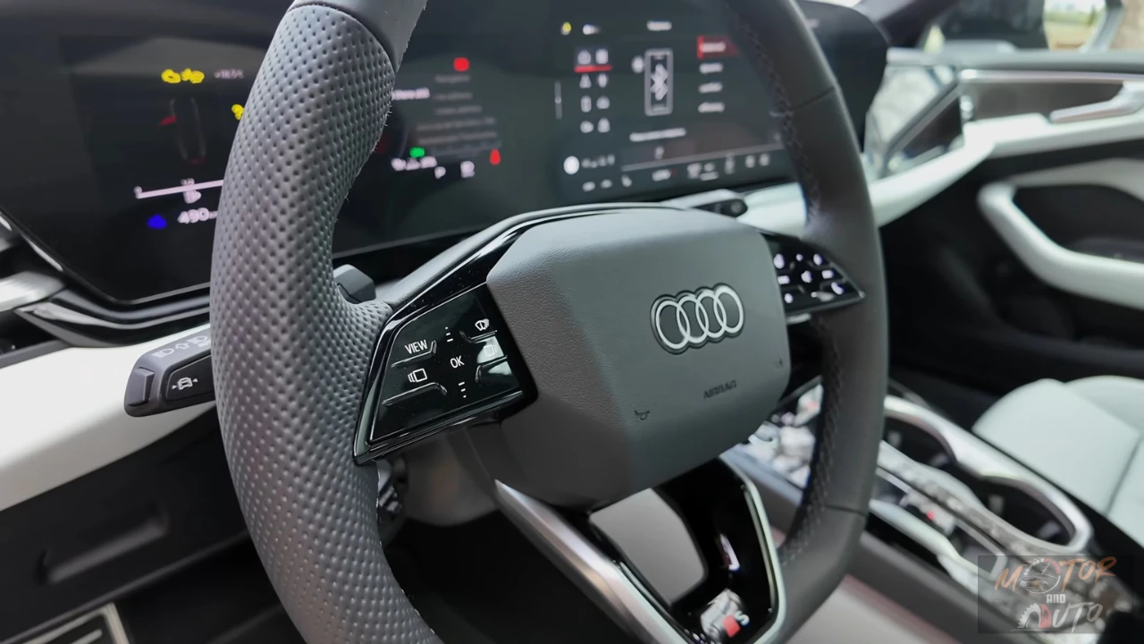 Gedetailleerd zicht op het stuurwiel van de 2026 Audi A6 met touch-gevoelige bedieningspanelen, Audi-logo, schakelpeddels en digitaal instrumentencluster op de achtergrond