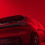 Achter driekwart aanzicht van 2026 Peugeot e-208 GTi met dakspoiler, GTi wielen, rode lakafwerking, en achterkant styling met prestatie modificaties