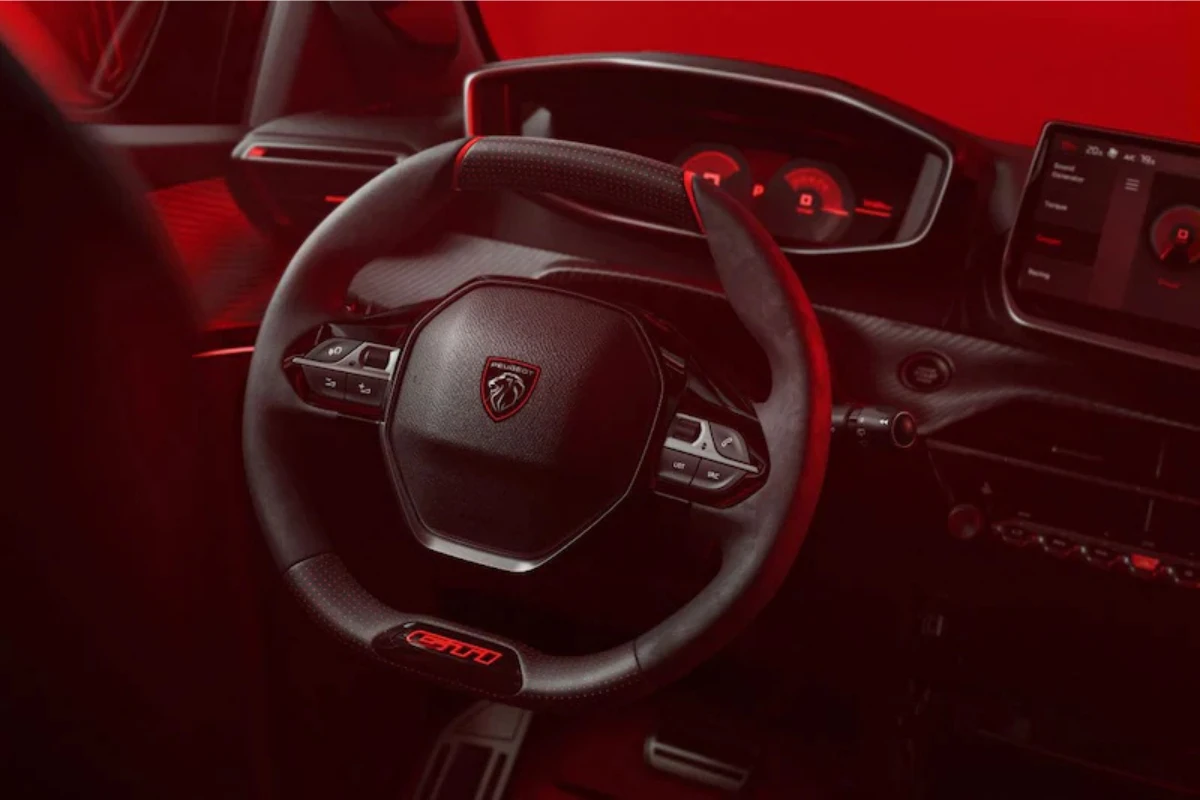 Interieur aanzicht van 2026 Peugeot e-208 GTi met dashboard, digitaal display met rode graphics, en GTi-gemerkt stuurwiel met rode accenten en geperforeerd leer