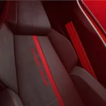 Close-up van 2026 Peugeot e-208 GTi voorstoel met rode centrale streep, gestructureerde stof, geïntegreerde hoofdsteun, en Peugeot leeuw logo op hoofdsteun
