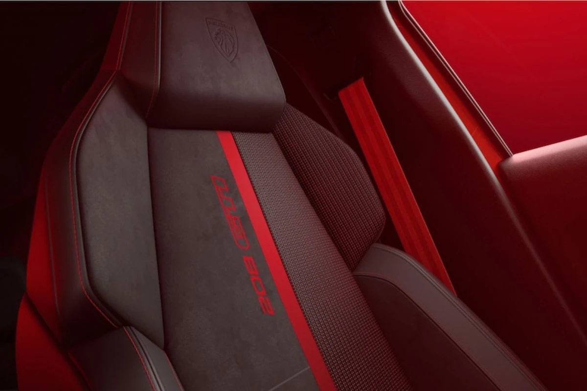 Close-up van 2026 Peugeot e-208 GTi voorstoel met rode centrale streep, gestructureerde stof, geïntegreerde hoofdsteun, en Peugeot leeuw logo op hoofdsteun