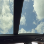 2026 Kia Sportage panoramadak met blauwe lucht en wolken door grote glas opening met zonnescherm en dak bediening zichtbaar