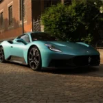 Voor driekwart aanzicht van turquoise 2026 Maserati MCPura op stedelijke kinderkoppenstraat met historisch bakstenen gebouw op achtergrond, met vernieuwd voorbumperontwerp en kenmerkende Maserati drietand grille
