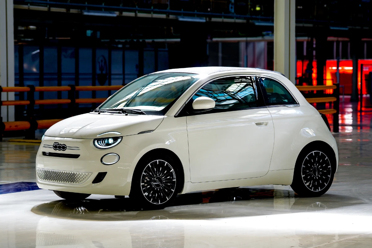 Witte Fiat 500 Hybrid in moderne fabriek omgeving met kleurrijke industriële verlichting en stalen raamwerk zichtbaar op de achtergrond