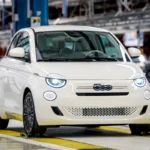Witte Fiat 500 Hybrid gepositioneerd op gele fabrieksvloer markeringen bij Mirafiori productiefaciliteit met industriële assemblage lijn achtergrond