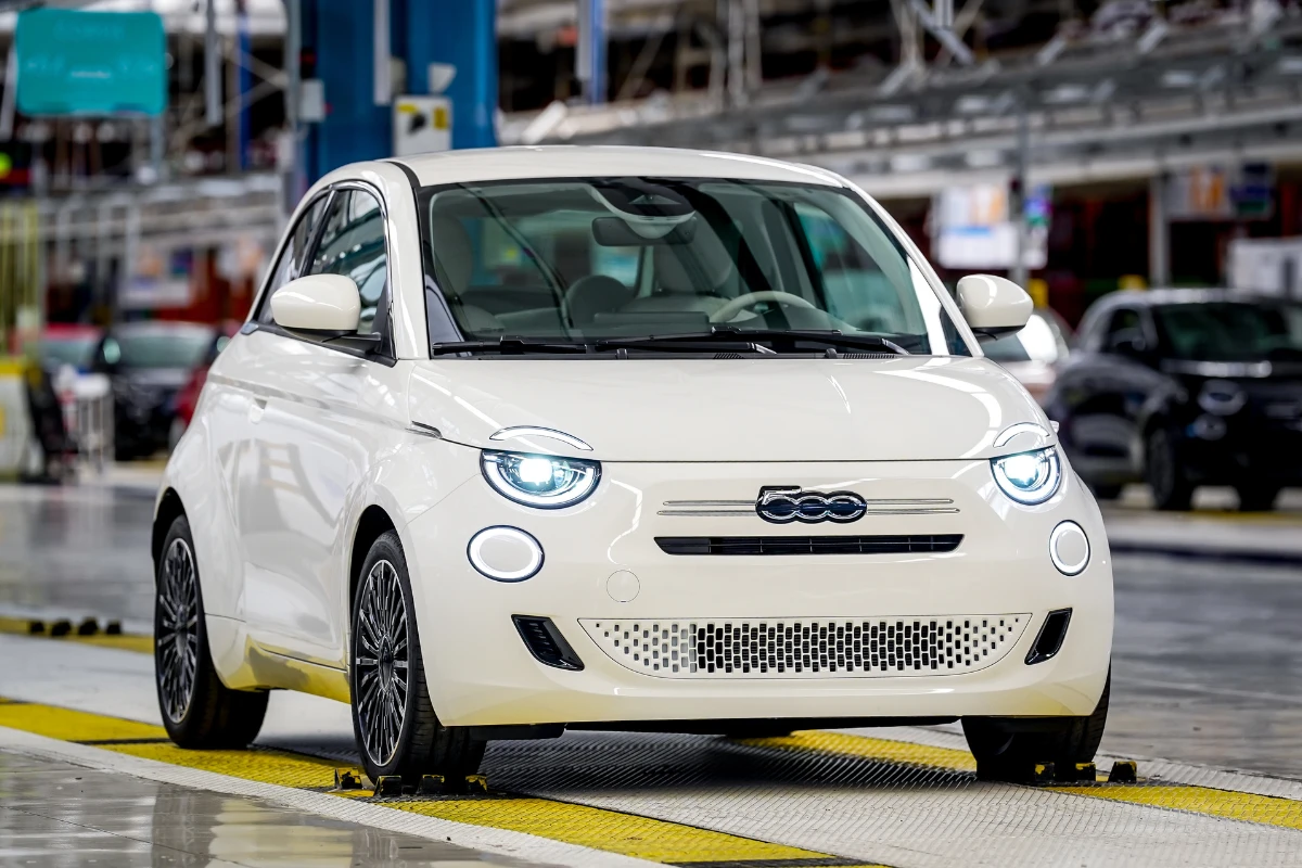 Witte Fiat 500 Hybrid gepositioneerd op gele fabrieksvloer markeringen bij Mirafiori productiefaciliteit met industriële assemblage lijn achtergrond