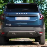 Direct achteraanzicht van blauwe 2025 Dacia Bigster SUV in bosomgeving met DACIA letters over de achterklep, BIGSTER modelbadge, Y-vormige LED achterlichten en achterbumperbescherming met parkeersensoren