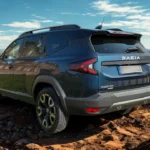Donkerblauwe 2025 Dacia Bigster SUV gefotografeerd vanuit de achter driekwart hoek in een rotsachtige woestijnomgeving met bergen op de achtergrond, waarbij de kenmerkende Y-vormige LED-achterlichten en BIGSTER-badge te zien zijn