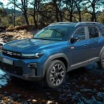 Blauwe 2025 Dacia Bigster SUV die een ondiepe rotsachtige beek oversteekt met water dat rond de banden spat, omringd door bomen en natuurlijk terrein, waarbij zijn off-road capaciteit en hoge bodemvrijheid wordt getoond