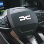 Stuurwiel van 2025 Dacia Bigster met zwarte afwerking en prominent DC-logo in het midden, bedieningsknoppen op spaken en gedeeltelijk zicht op digitaal instrumentenpaneel