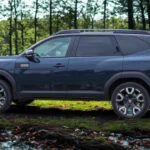 Volledig zijaanzicht van blauwe 2025 Dacia Bigster SUV in bosomgeving waarbij het volledige voertuigprofiel van voor naar achter te zien is, met nadruk op de verlengde wielbasis, dakrails en beschermende zijbekleding