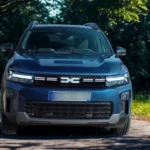 Blauwe 2025 Dacia Bigster SUV voorwaarts gericht op een met bomen omzoomde weg met verlichte LED-koplampen, met de prominente DC-logo grille en Y-vormige DRL-signatuur in een natuurlijke bosomgeving