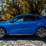 2025 Ford Kuga in opvallende blauwe metallic lak op Europese kinderkopjesstraat, elegante zijprofiel met vloeiende lijnen en moderne SUV-proporties tonend