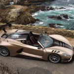 Hennessey Venom F5 Revolution LF Roadster met dak verwijderd gepositioneerd op kustweg met oceaan en rotsachtige kliffen op achtergrond, champagne metallic lak tonend