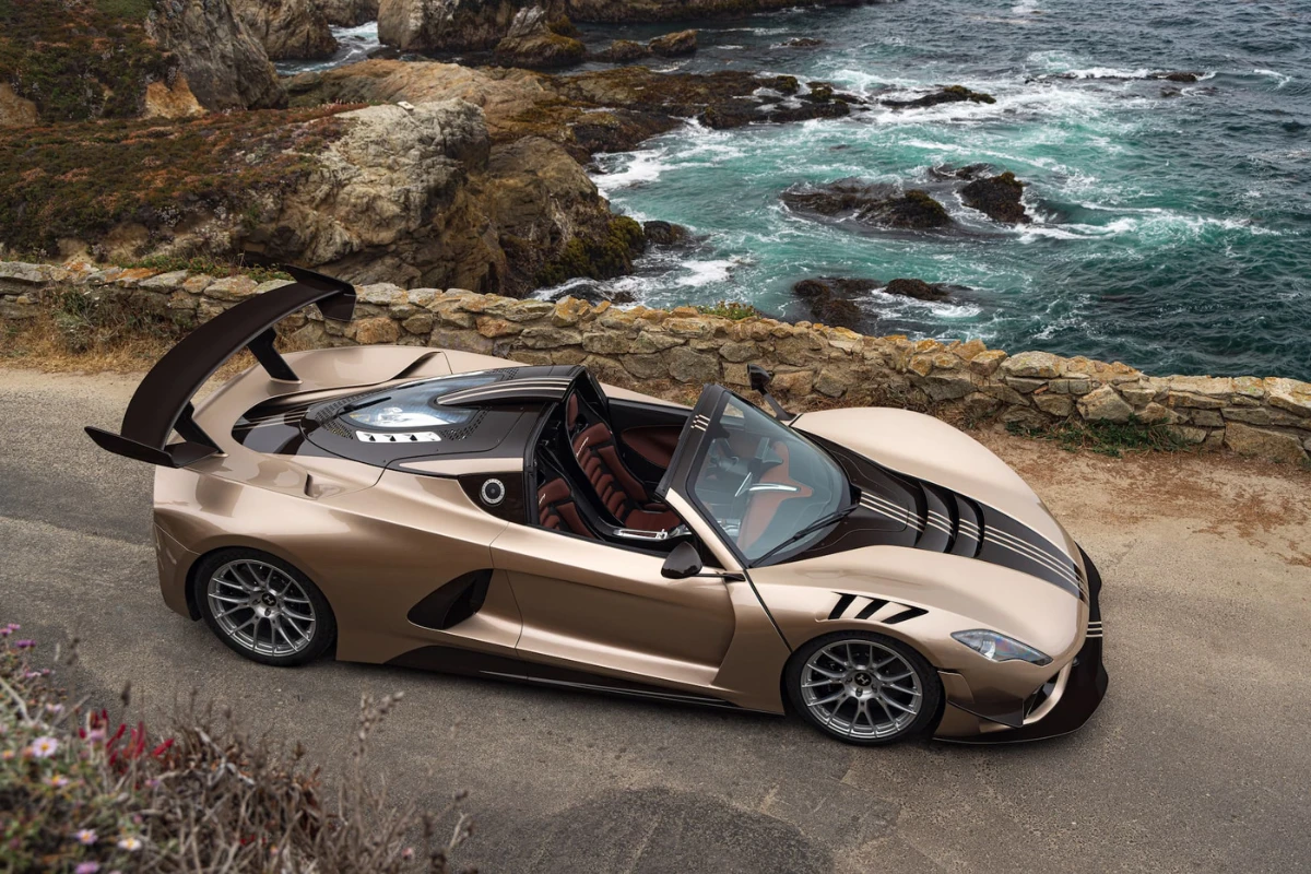 Hennessey Venom F5 Revolution LF Roadster met dak verwijderd gepositioneerd op kustweg met oceaan en rotsachtige kliffen op achtergrond, champagne metallic lak tonend